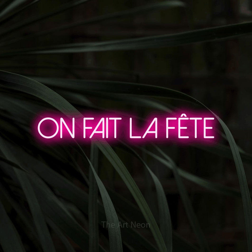 “On fait la fête” LED neon sign in hot pink, with thin sans-serif lettering, shown against a close-up of dark foliage.