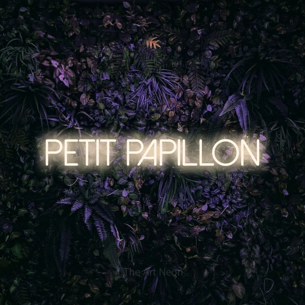 “Petit papillon” LED neon sign in warm white, with thin sans-serif lettering, shown against a dark wall with purple flowers.