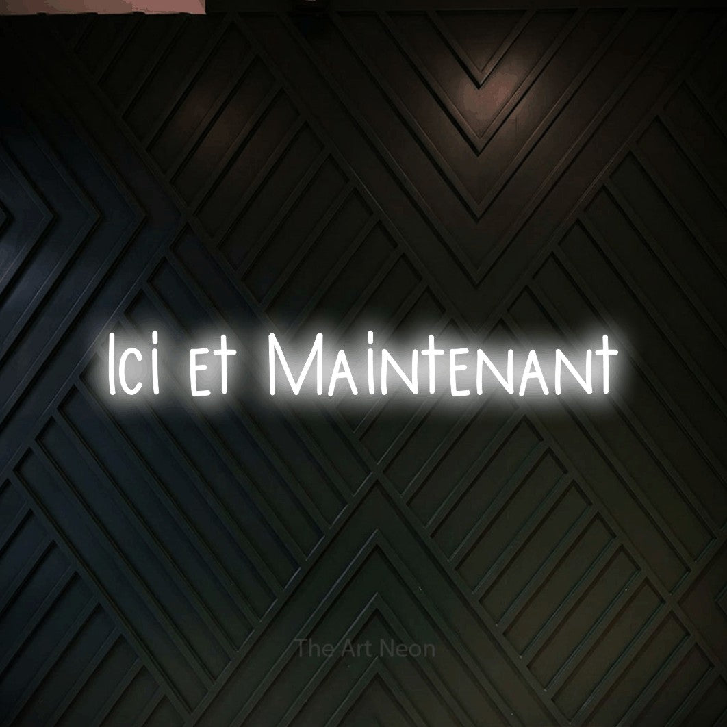 “Ici et maintenant” LED neon sign in cool white, with narrow hand-drawn lettering, shown against a black chevron-patterned wood wall.