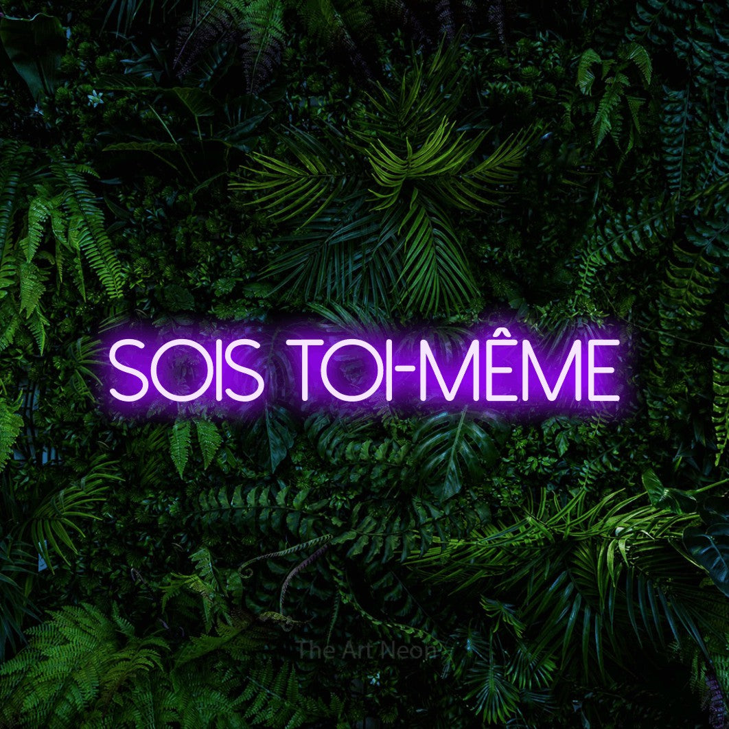 “Sois toi-même” LED neon sign in purple, with thin sans-serif lettering, shown against dense tropical foliage.