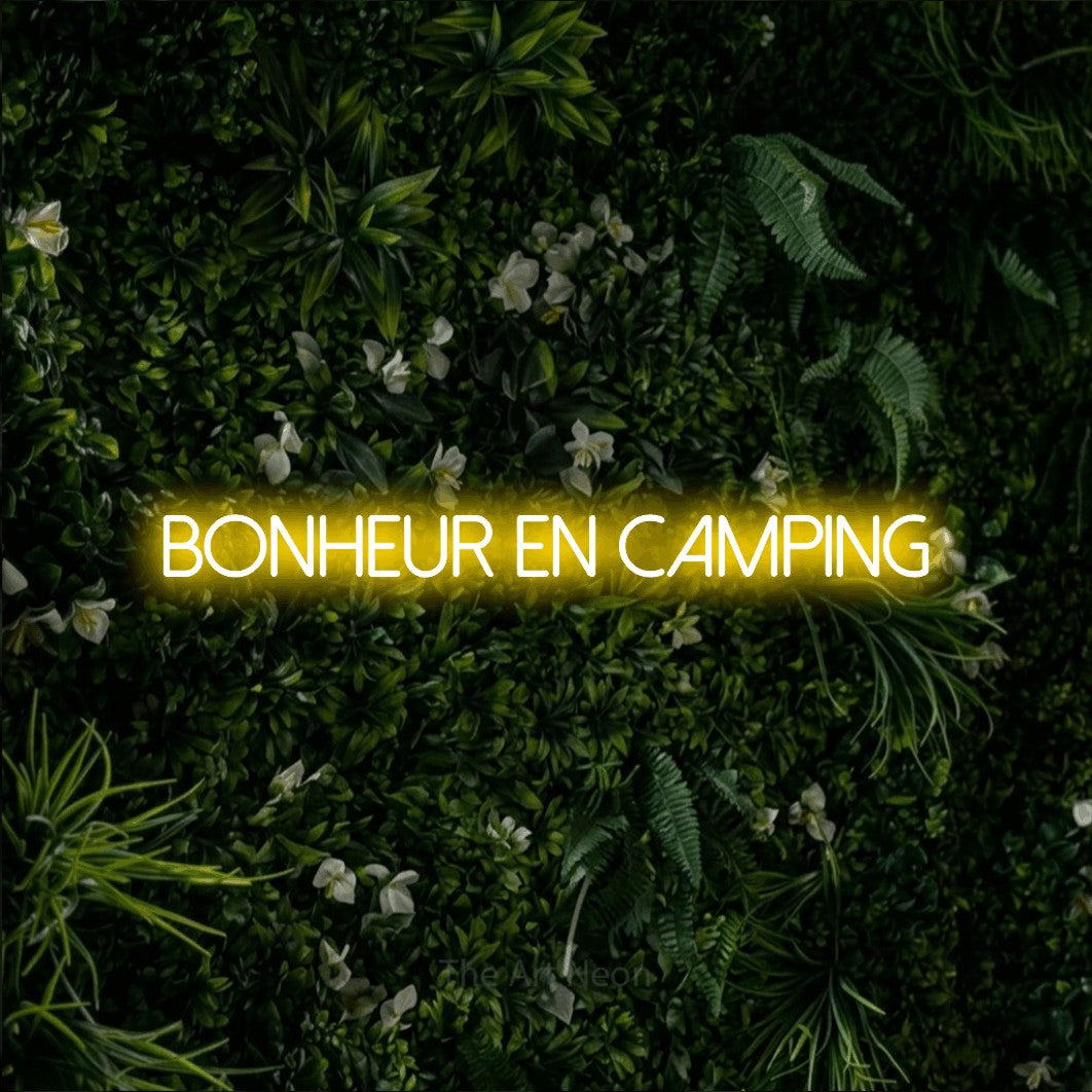 “Bonheur en camping” LED neon sign in yellow, with thin sans-serif lettering, shown against a dense green plant wall.