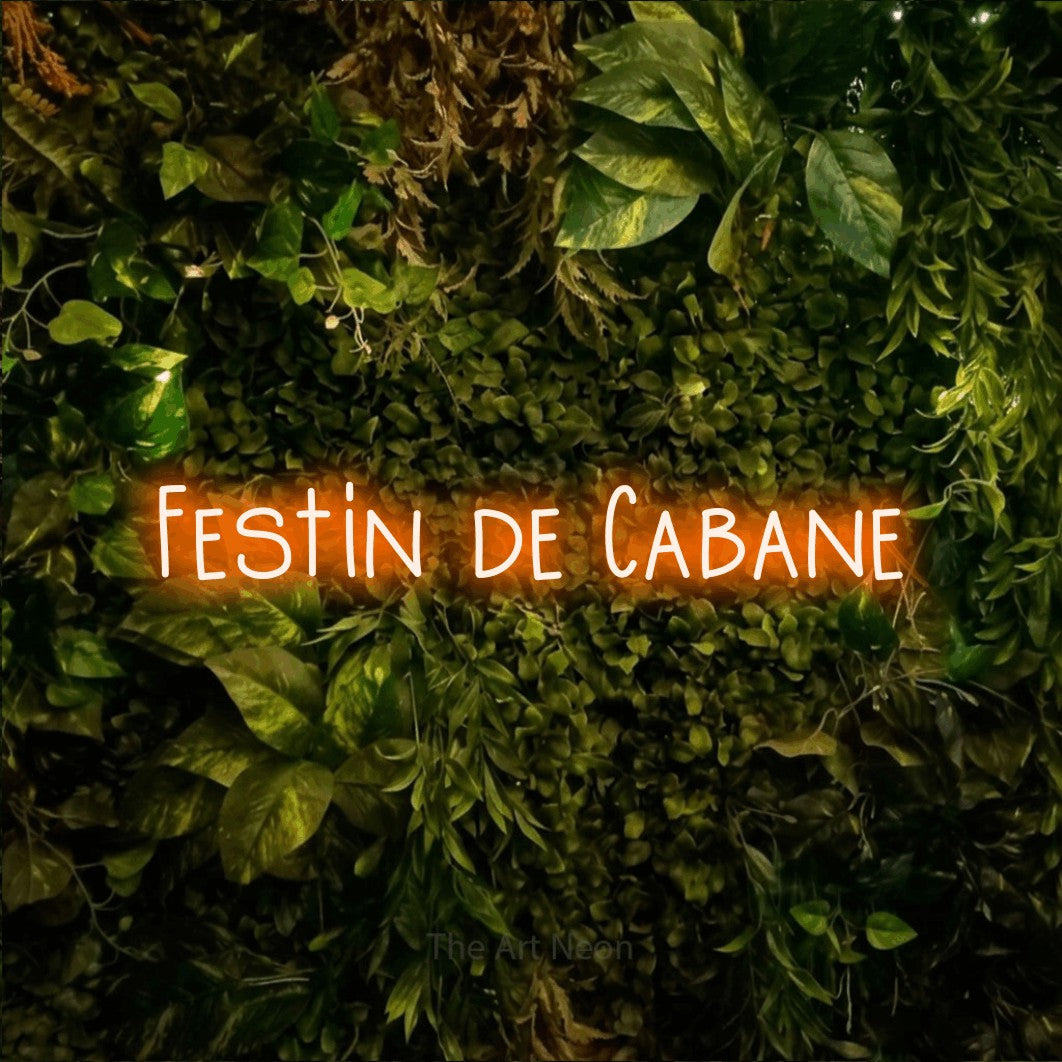 “Festin de cabane” LED neon sign in orange, with narrow hand-drawn lettering, shown against bright green and yellow foliage.