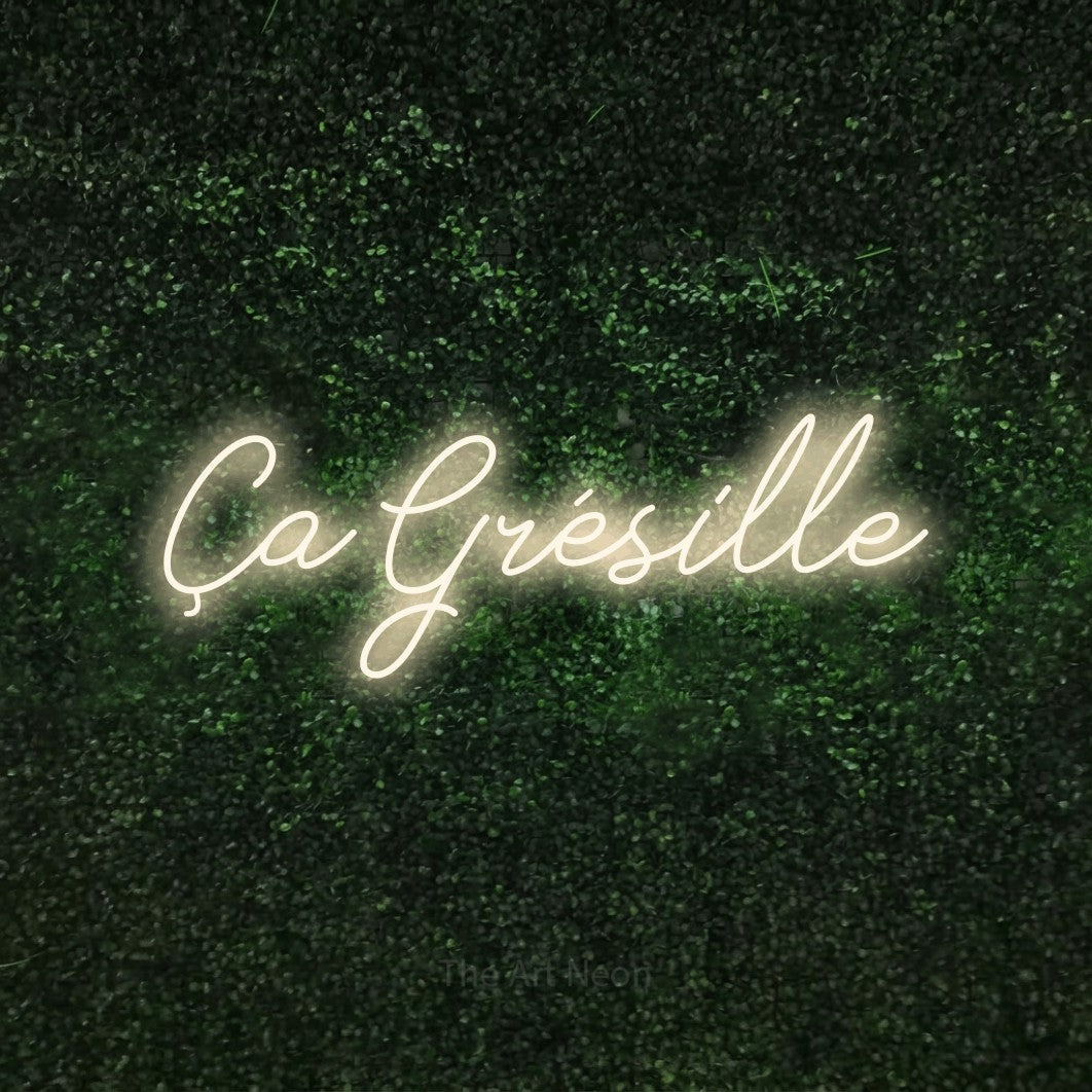 “Ça grésille” LED neon sign in warm white, with cursive lettering, shown against a textured green hedge.