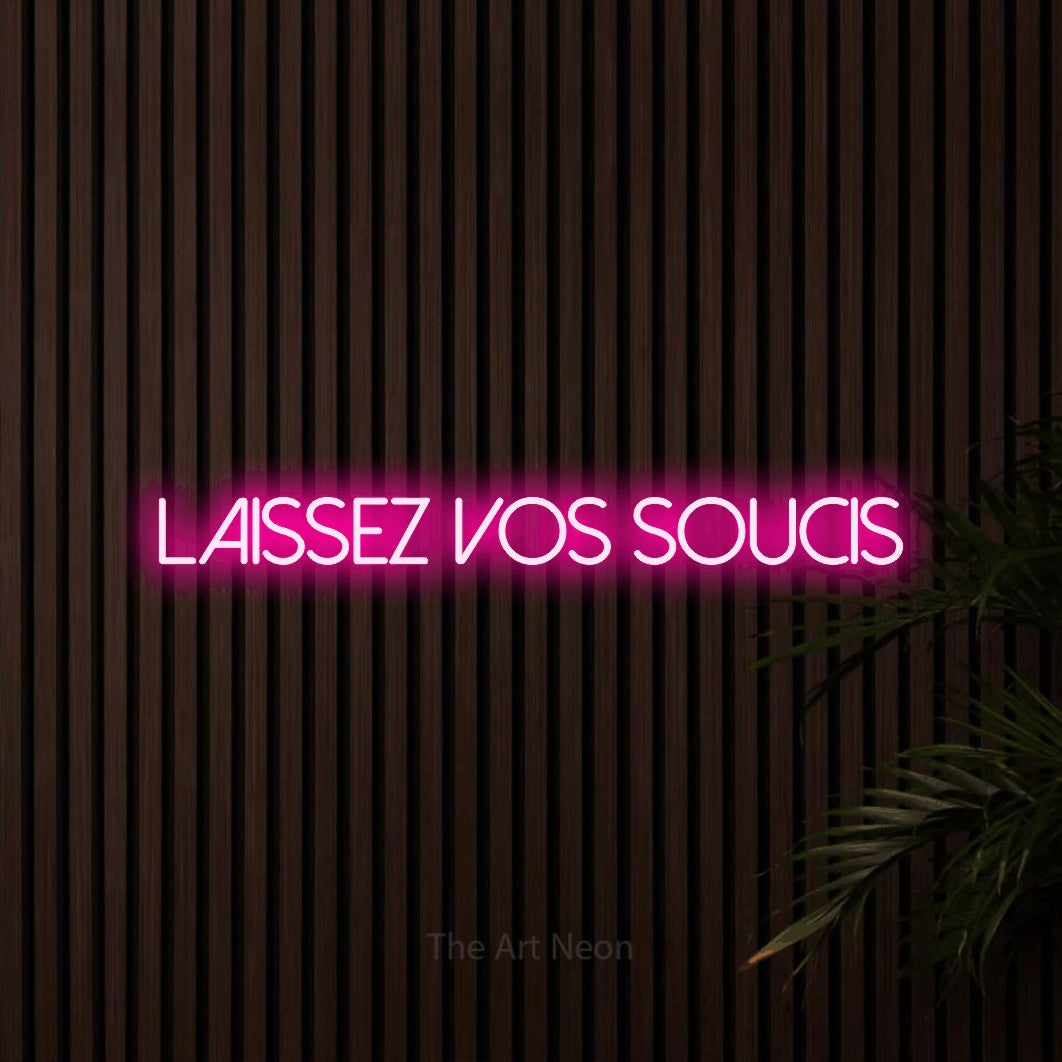 “Laissez vos soucis” LED neon sign in hot pink, with thin sans-serif lettering, shown against dark wooden wall slats.