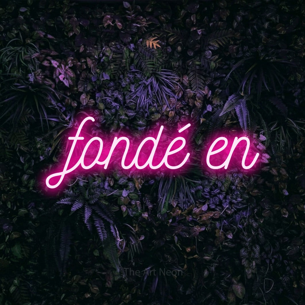 “Fondé en” LED neon sign in hot pink, with ornate cursive lettering, shown against a dark wall with purple flowers.