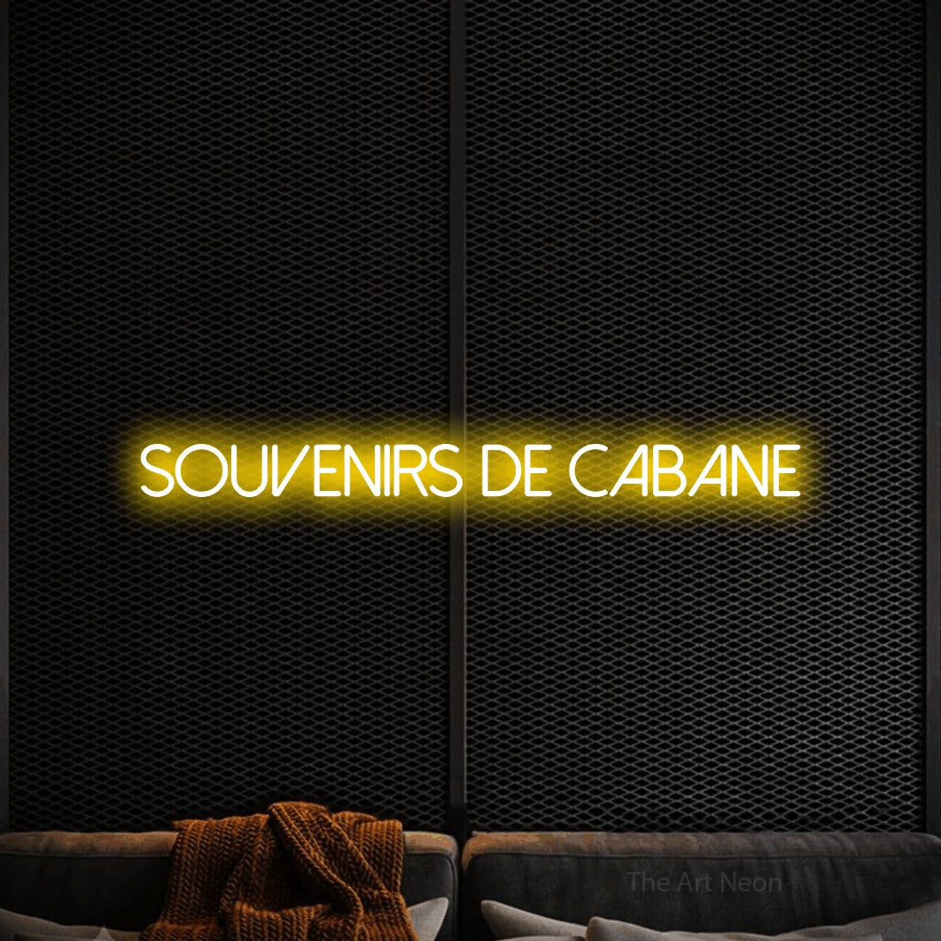“Souvenirs de cabane” LED neon sign in yellow, with thin sans-serif lettering, shown against a modern dark-toned seating area.