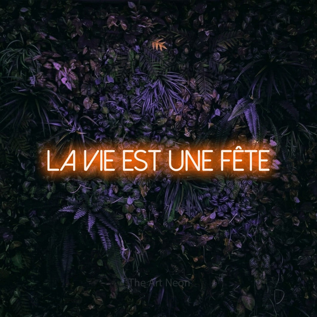“La vie est une fête” LED neon sign in orange, with thin sans-serif lettering, shown against a dark wall with purple flowers.