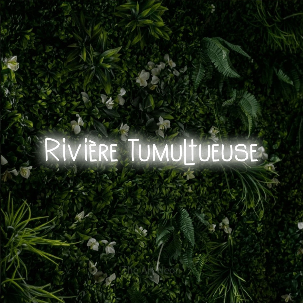 “Rivière tumultueuse” LED neon sign in cool white, with narrow hand-drawn lettering, shown against a dense green plant wall.