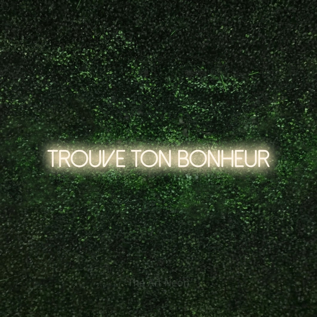 “Trouve ton bonheur” LED neon sign in warm white, with thin sans-serif lettering, shown against a textured green hedge.