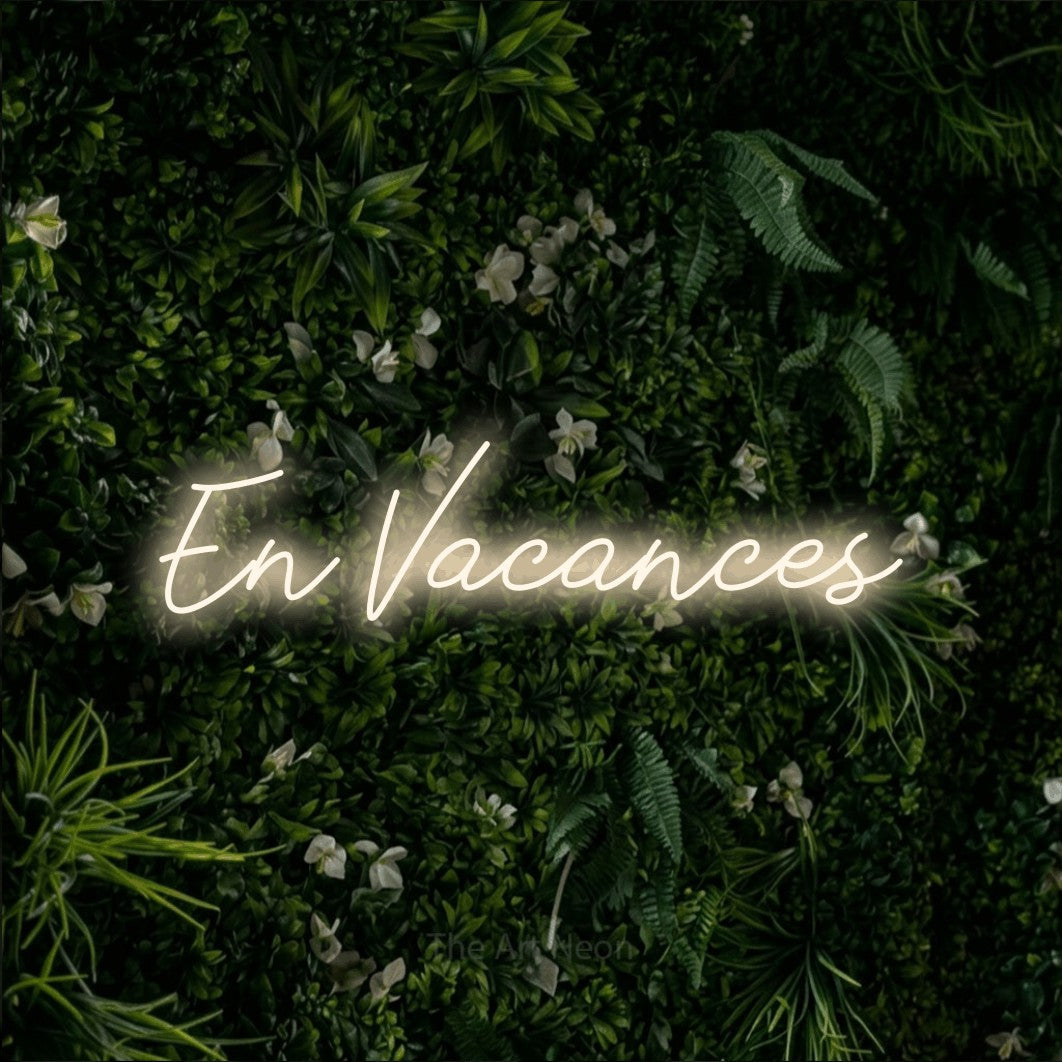 “En vacances” LED neon sign in warm white, with cursive lettering, shown against a dense green plant wall.