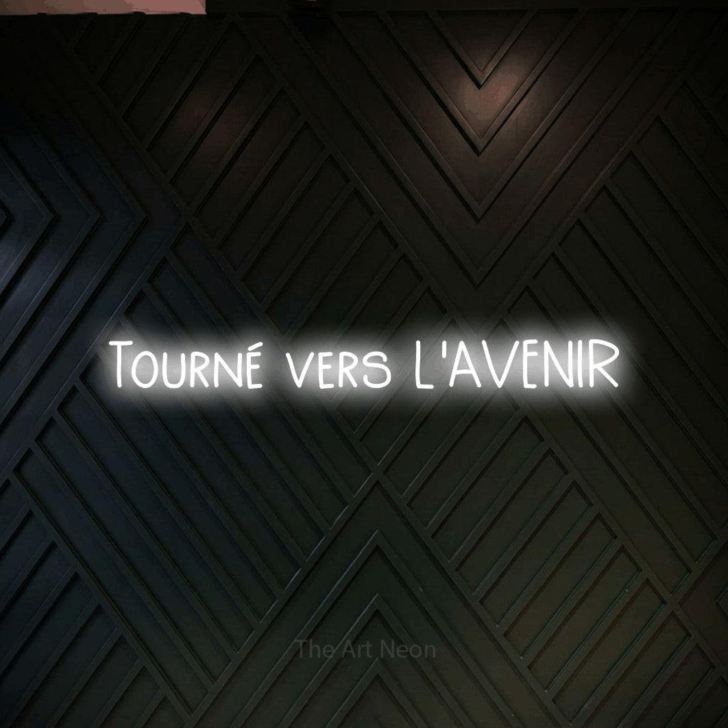 “Tourné vers l'avenir” LED neon sign in cool white, with narrow hand-drawn lettering, shown against a black chevron-patterned wood wall.