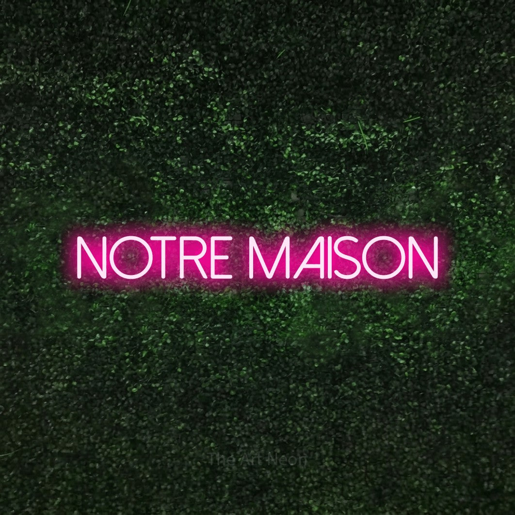 Aperçu d’une enseigne néon LED « Notre maison » en rose vif, en lettres fines sans empattement, photographiée sur une haie verte texturée, pour un salon.