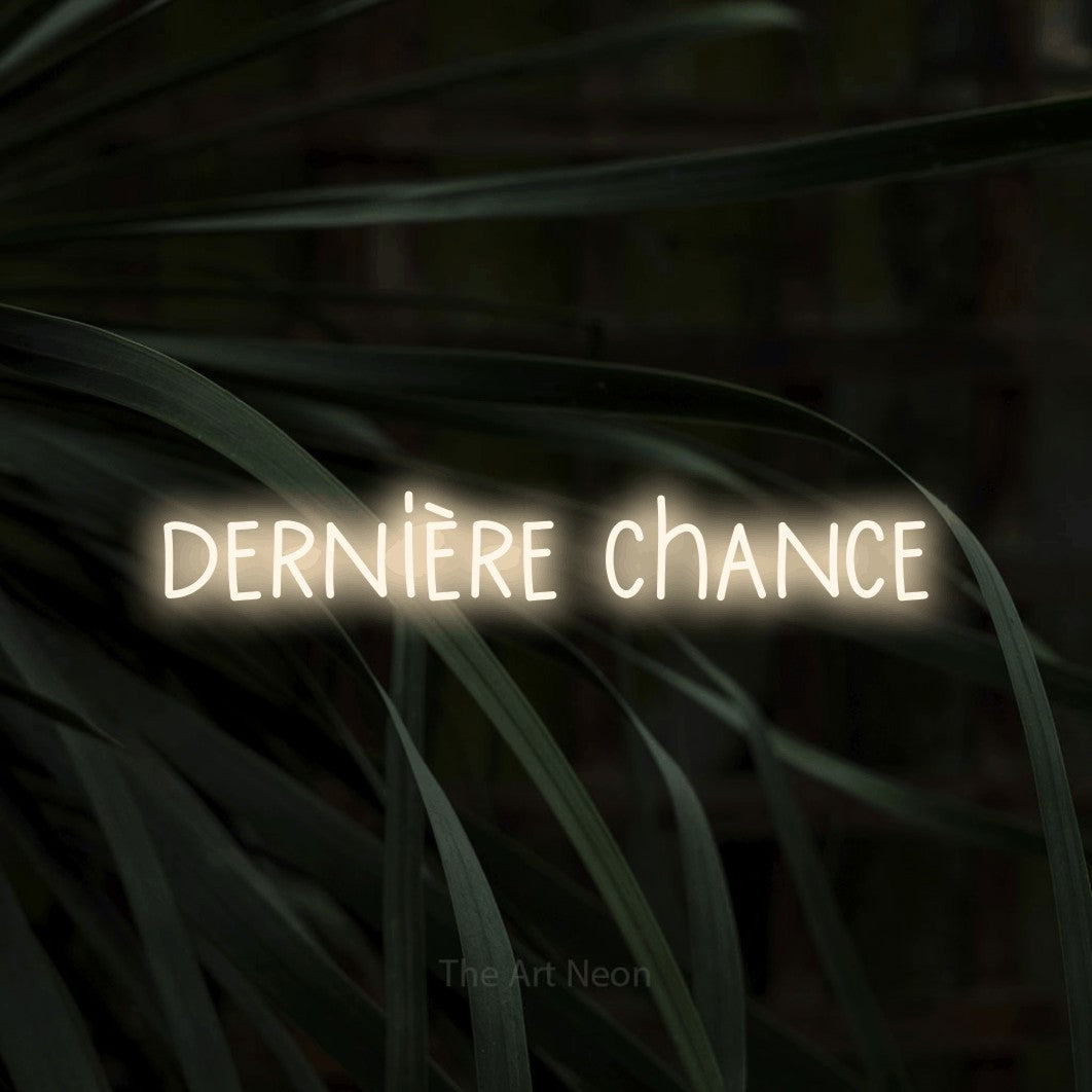 “Dernière chance” LED neon sign in warm white, with narrow hand-drawn lettering, shown against a close-up of dark foliage.
