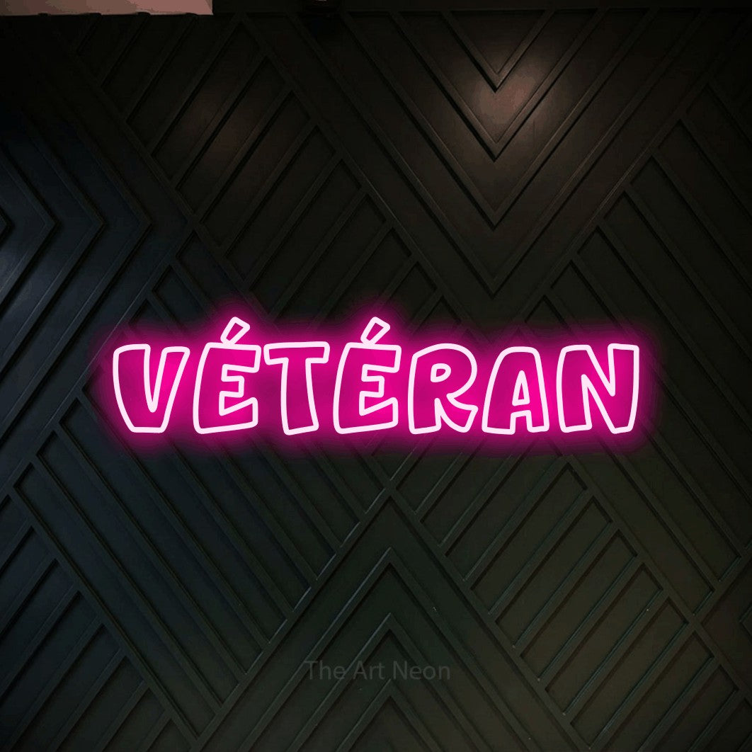 “Vétéran” LED neon sign in hot pink, with rounded outline lettering, shown against a black chevron-patterned wood wall.