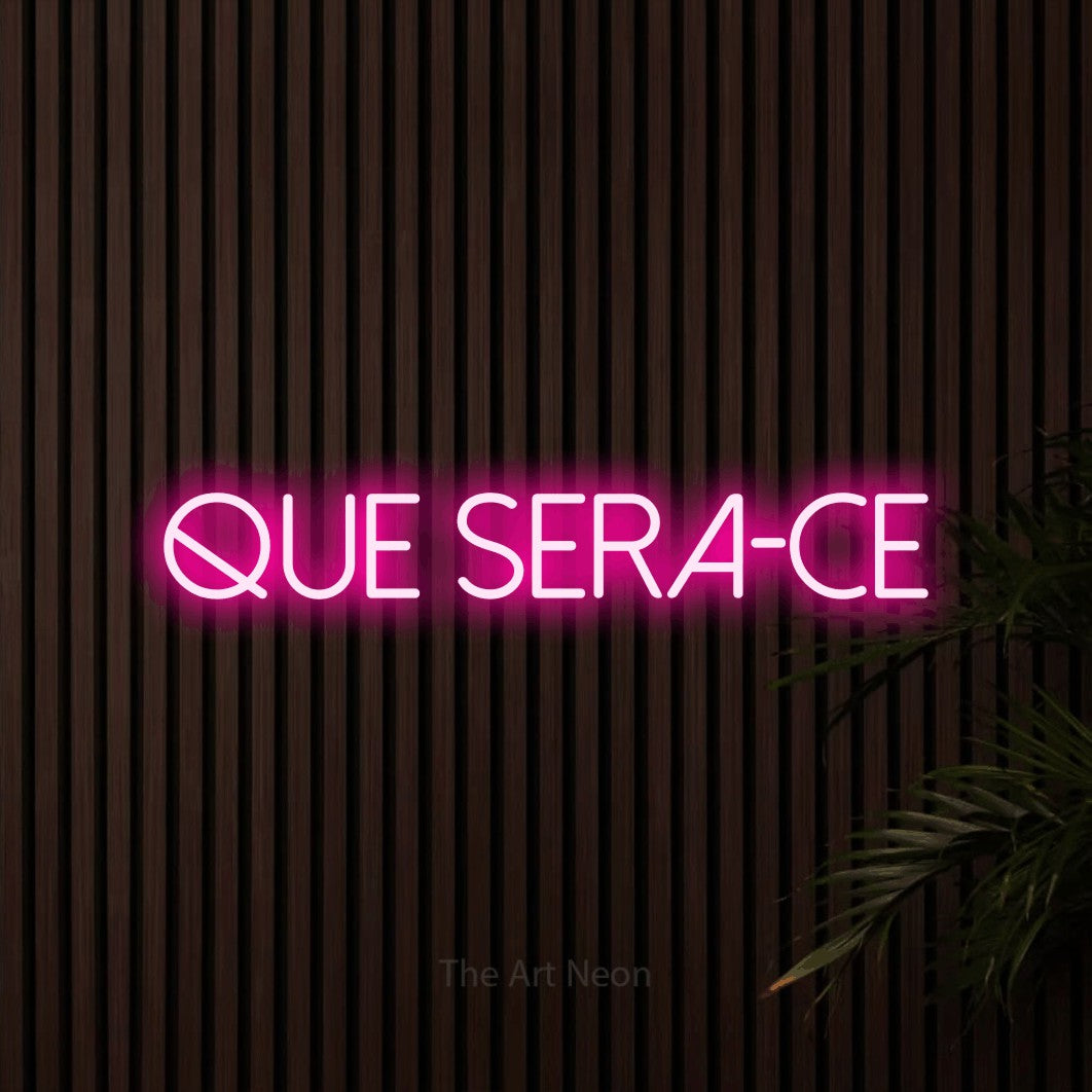 “Que sera-ce” LED neon sign in hot pink, with thin sans-serif lettering, shown against dark wooden wall slats.