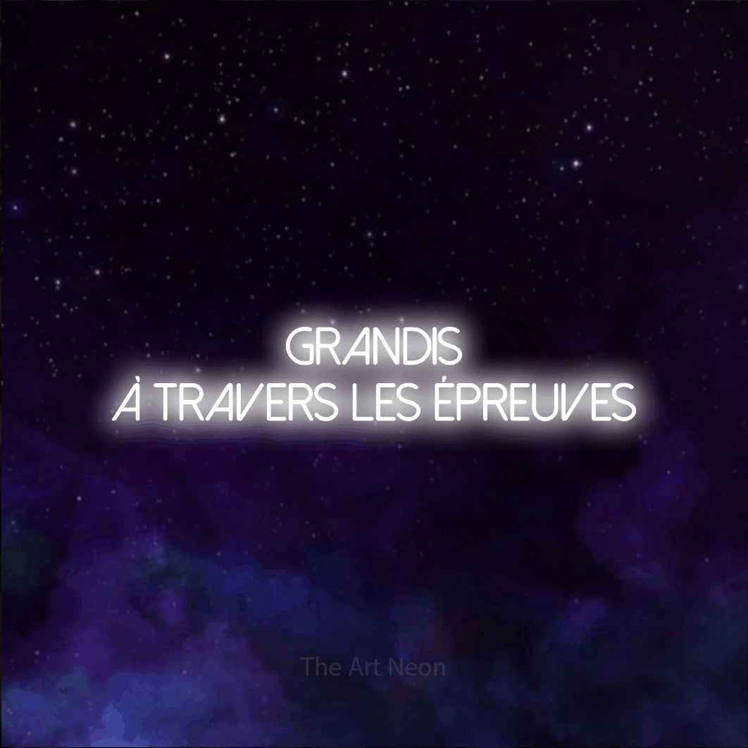 “Grandis à travers les épreuves” LED neon sign in cool white, with thin sans-serif lettering across 2 lines, shown against a purple galaxy backdrop.