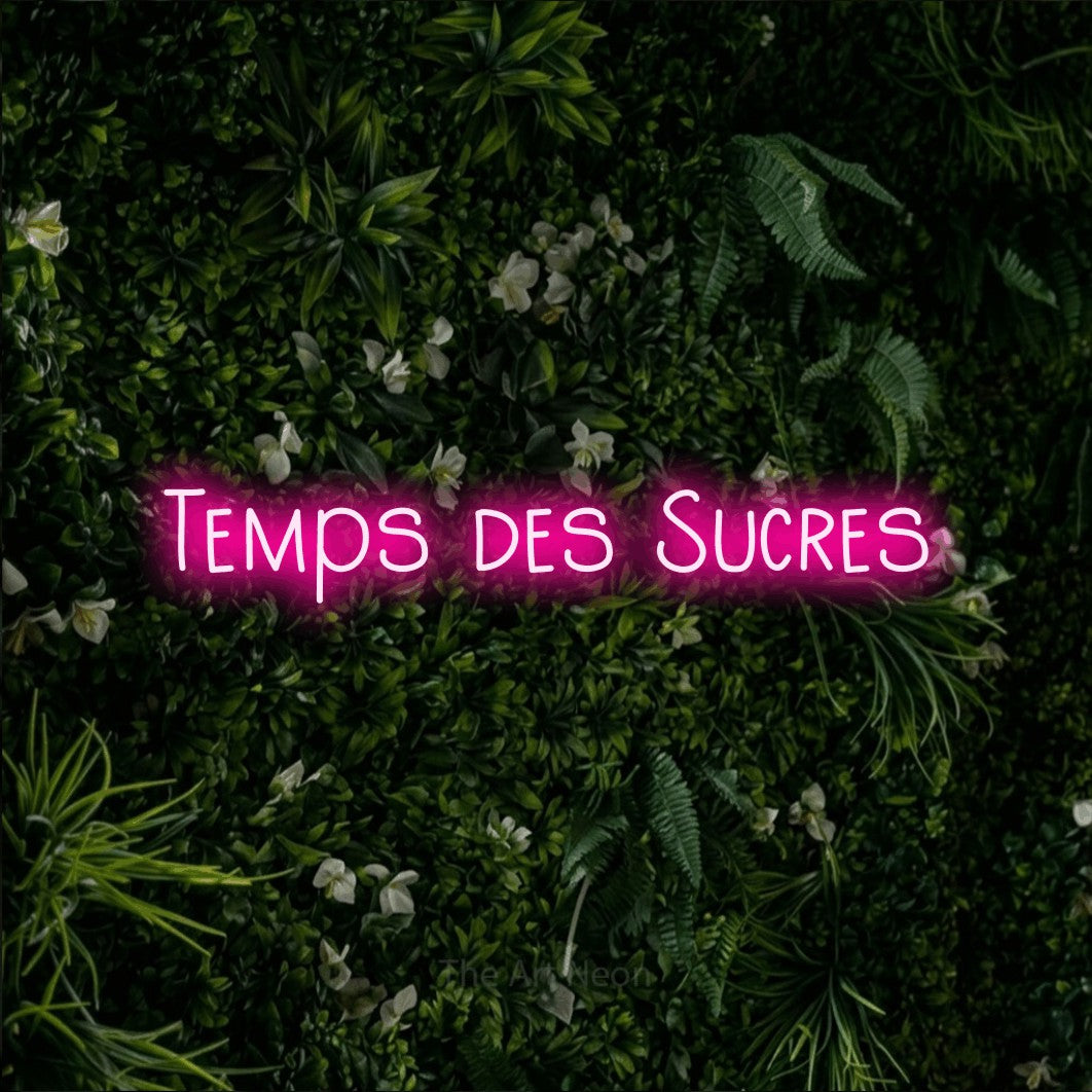 “Temps des sucres” LED neon sign in hot pink, with narrow hand-drawn lettering, shown against a dense green plant wall.