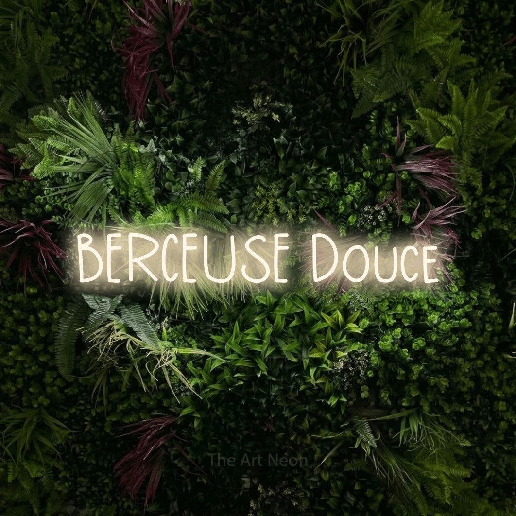 “Berceuse douce” LED neon sign in warm white, with narrow hand-drawn lettering, shown against a plant wall with pink flowers.