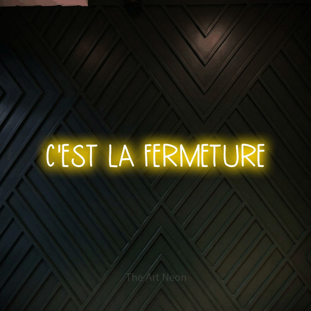 “C'est la fermeture” LED neon sign in yellow, with narrow hand-drawn lettering, shown against a black chevron-patterned wood wall.