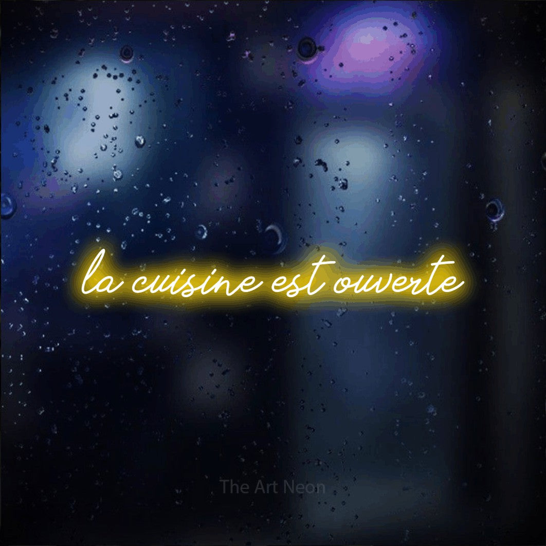“La cuisine est ouverte” LED neon sign in yellow, with cursive lettering, shown against glass with blue and purple reflections.