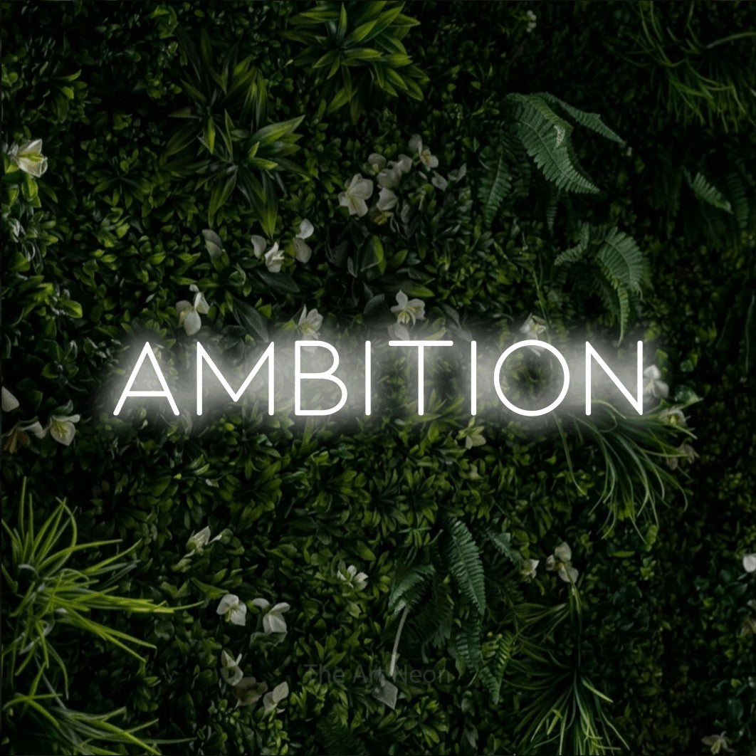 “Ambition” LED neon sign in cool white, with thin, widely spaced lettering, shown against a dense green plant wall.