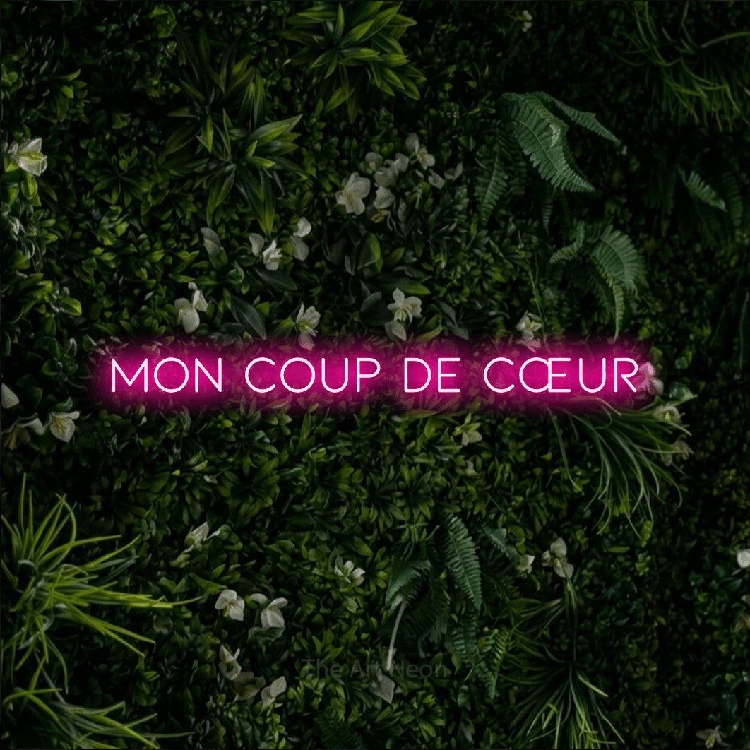 “Mon coup de cœur” LED neon sign in hot pink, with thin, widely spaced lettering, shown against a dense green plant wall.