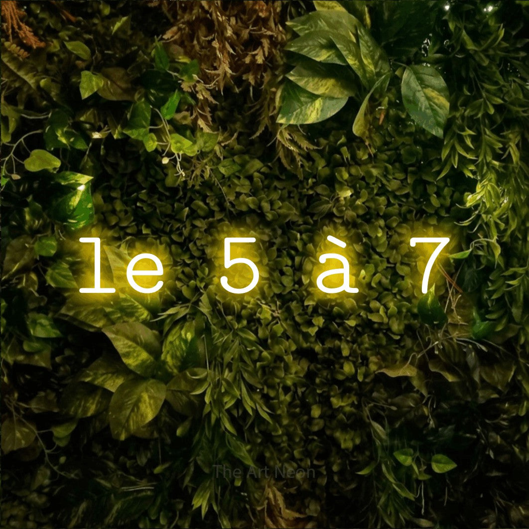 “Le 5 à 7” LED neon sign in yellow, with typewriter-style lettering, shown against bright green and yellow foliage.