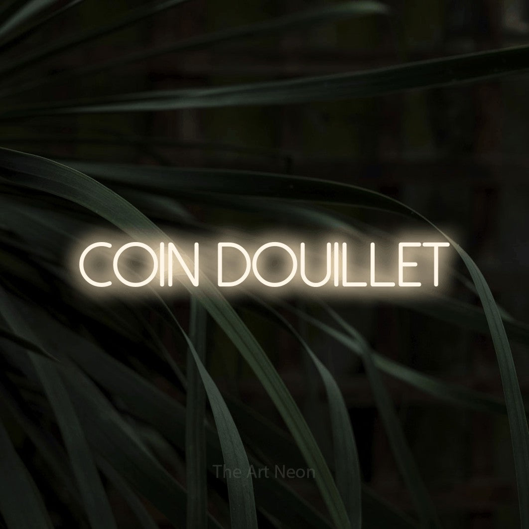 “Coin douillet” LED neon sign in warm white, with thin sans-serif lettering, shown against a close-up of dark foliage.