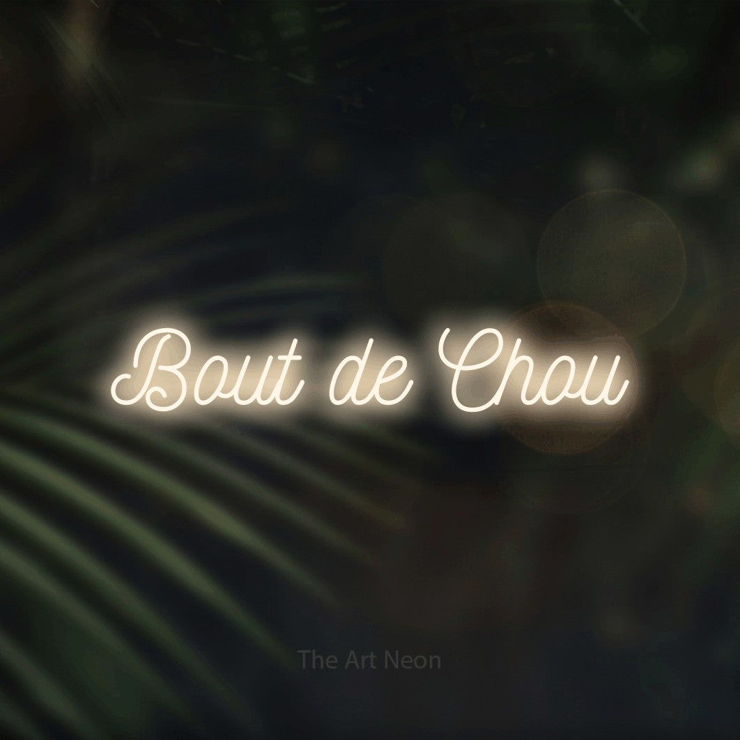 “Bout de chou” LED neon sign in warm white, with ornate cursive lettering, shown against a blurred palm and bokeh-light backdrop.