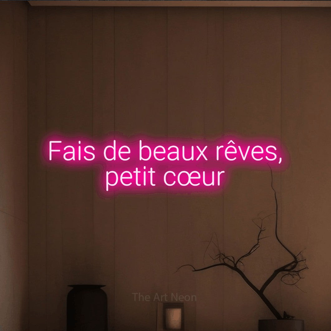 “Fais de beaux rêves, petit cœur” LED neon sign in hot pink, with rounded sans-serif lettering across 2 lines, shown against a simple earth-toned interior.