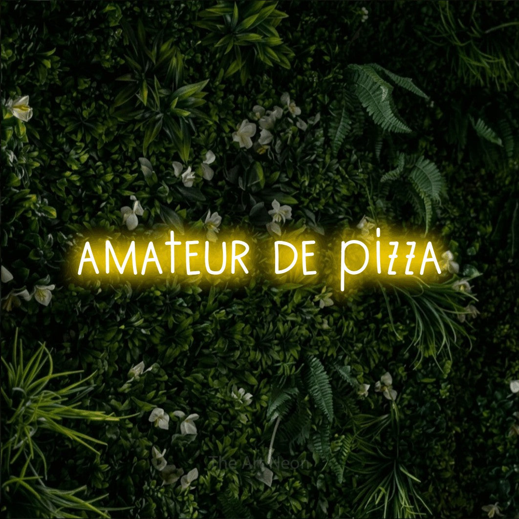 “Amateur de pizza” LED neon sign in yellow, with narrow hand-drawn lettering, shown against a dense green plant wall.