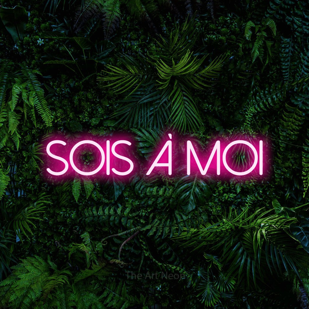 “Sois à moi” LED neon sign in hot pink, with thin sans-serif lettering, shown against dense tropical foliage.