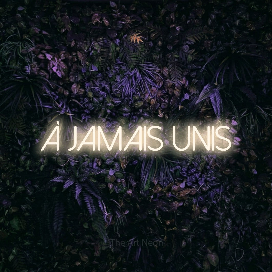 “À jamais unis” LED neon sign in warm white, with thin sans-serif lettering, shown against a dark wall with purple flowers.