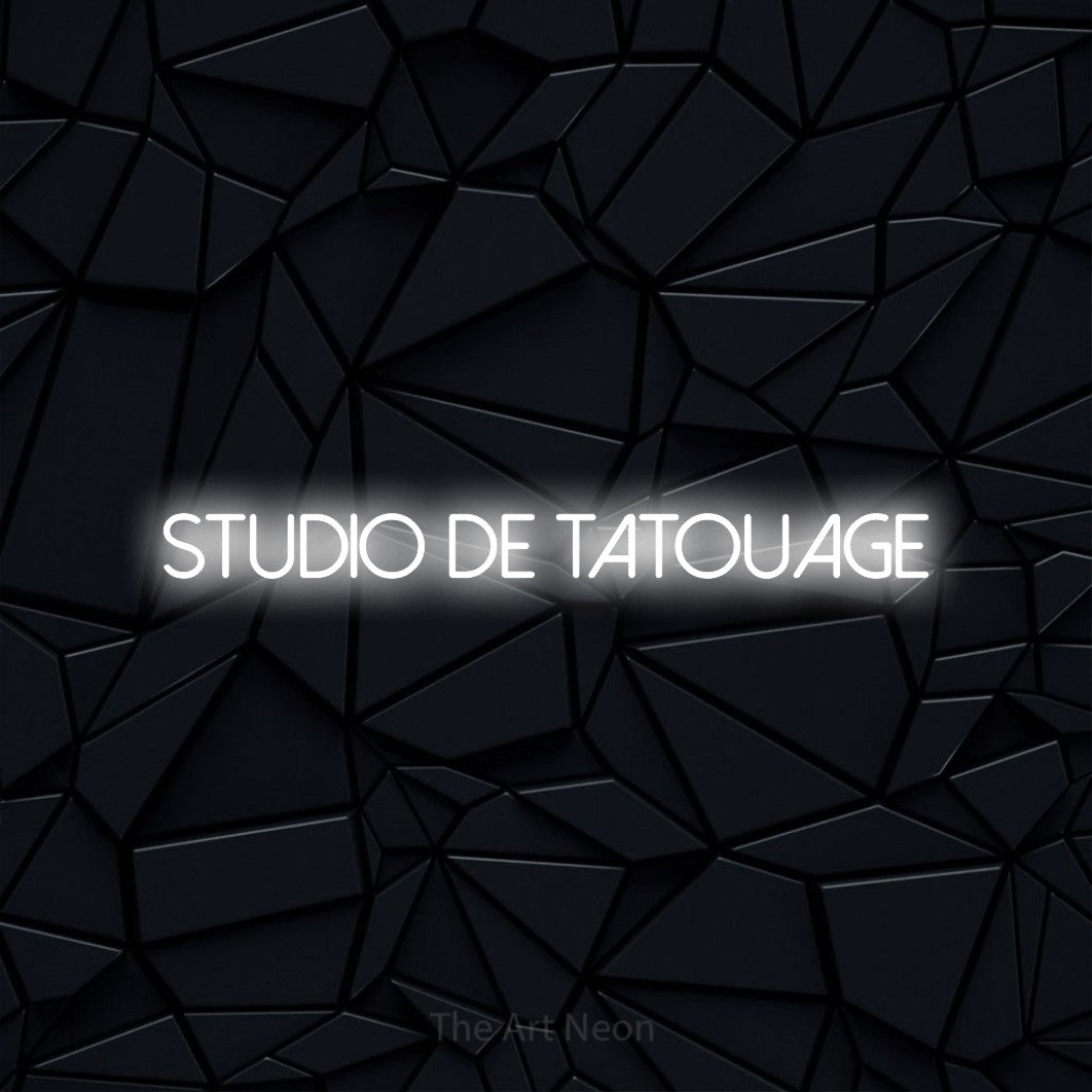 “Studio de tatouage” LED neon sign in cool white, with thin sans-serif lettering, shown against a black polygonal surface.