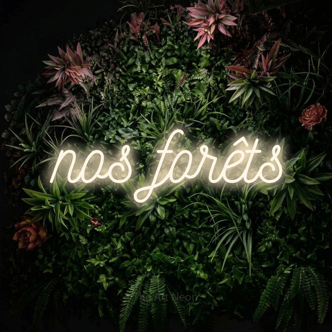 “Nos forêts” LED neon sign in warm white, with ornate cursive lettering, shown against a flowering plant wall.
