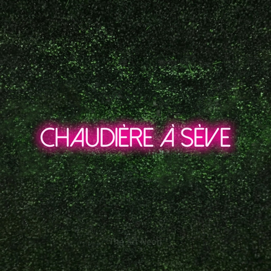 “Chaudière à sève” LED neon sign in hot pink, with thin sans-serif lettering, shown against a textured green hedge.