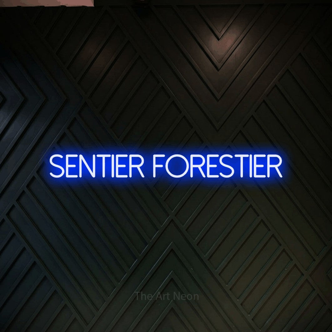 “Sentier forestier” LED neon sign in blue, with thin sans-serif lettering, shown against a black chevron-patterned wood wall.