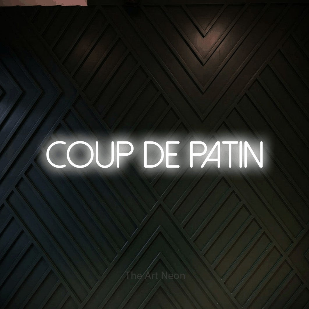 “Coup de patin” LED neon sign in cool white, with thin sans-serif lettering, shown against a black chevron-patterned wood wall.