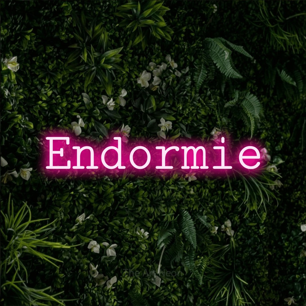 “Endormie” LED neon sign in hot pink, with typewriter-style lettering, shown against a dense green plant wall.
