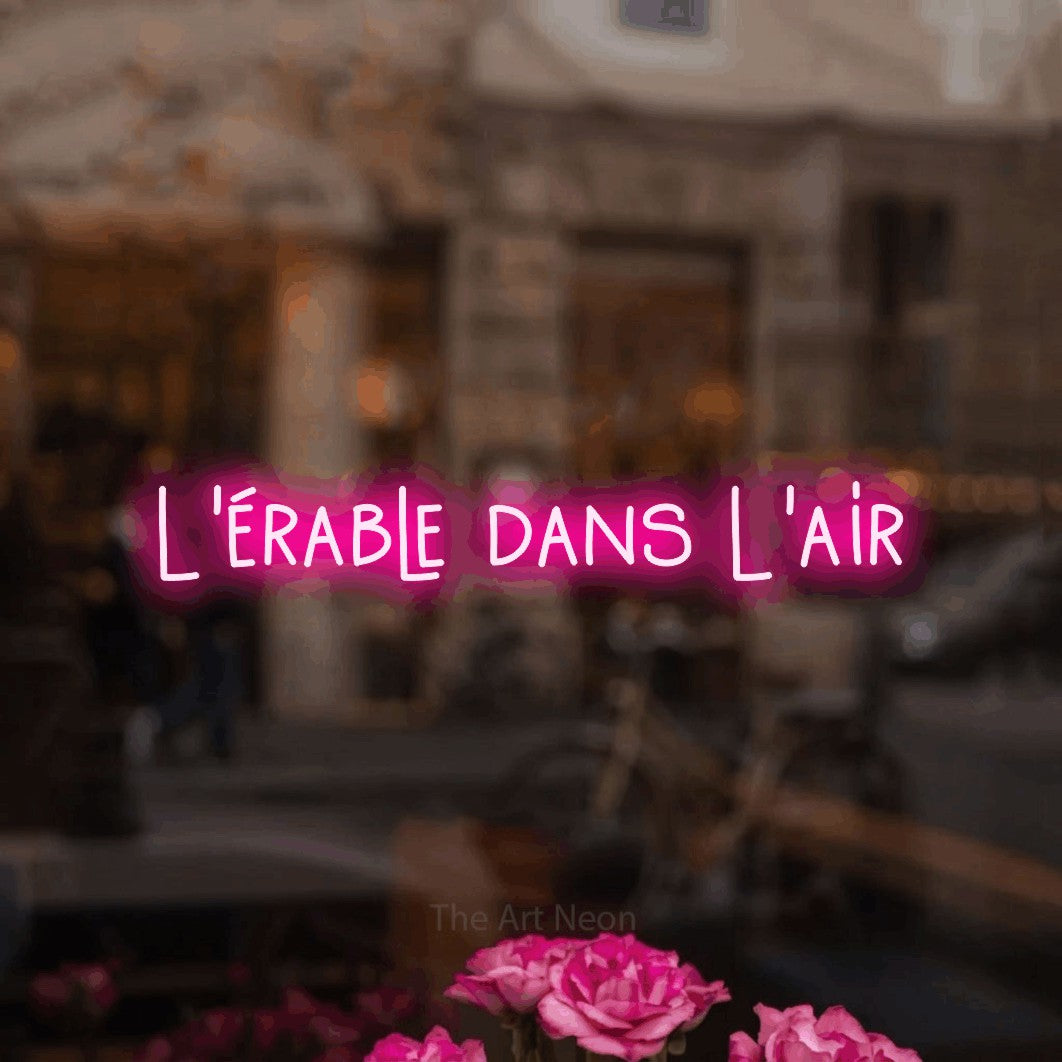 “L'érable dans l'air” LED neon sign in hot pink, with narrow hand-drawn lettering, shown against a blurred café-bar interior.