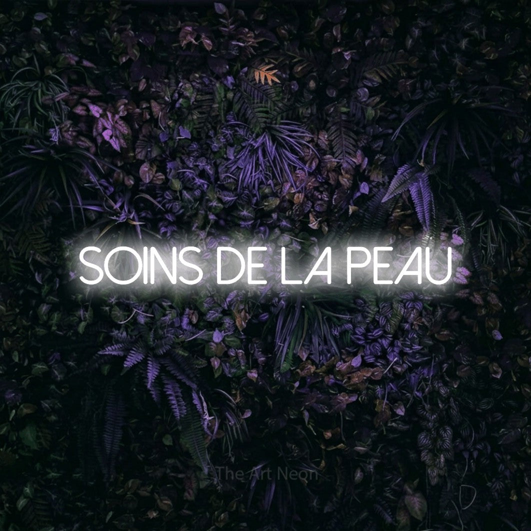 Aperçu d’une enseigne néon LED « Soins de la peau » en blanc froid, en lettres fines sans empattement, photographiée sur un mur sombre aux fleurs violettes, pour un salon de beauté.