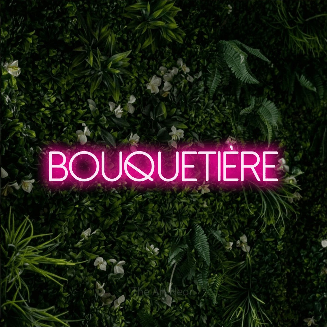 “Bouquetière” LED neon sign in hot pink, with thin sans-serif lettering, shown against a dense green plant wall.