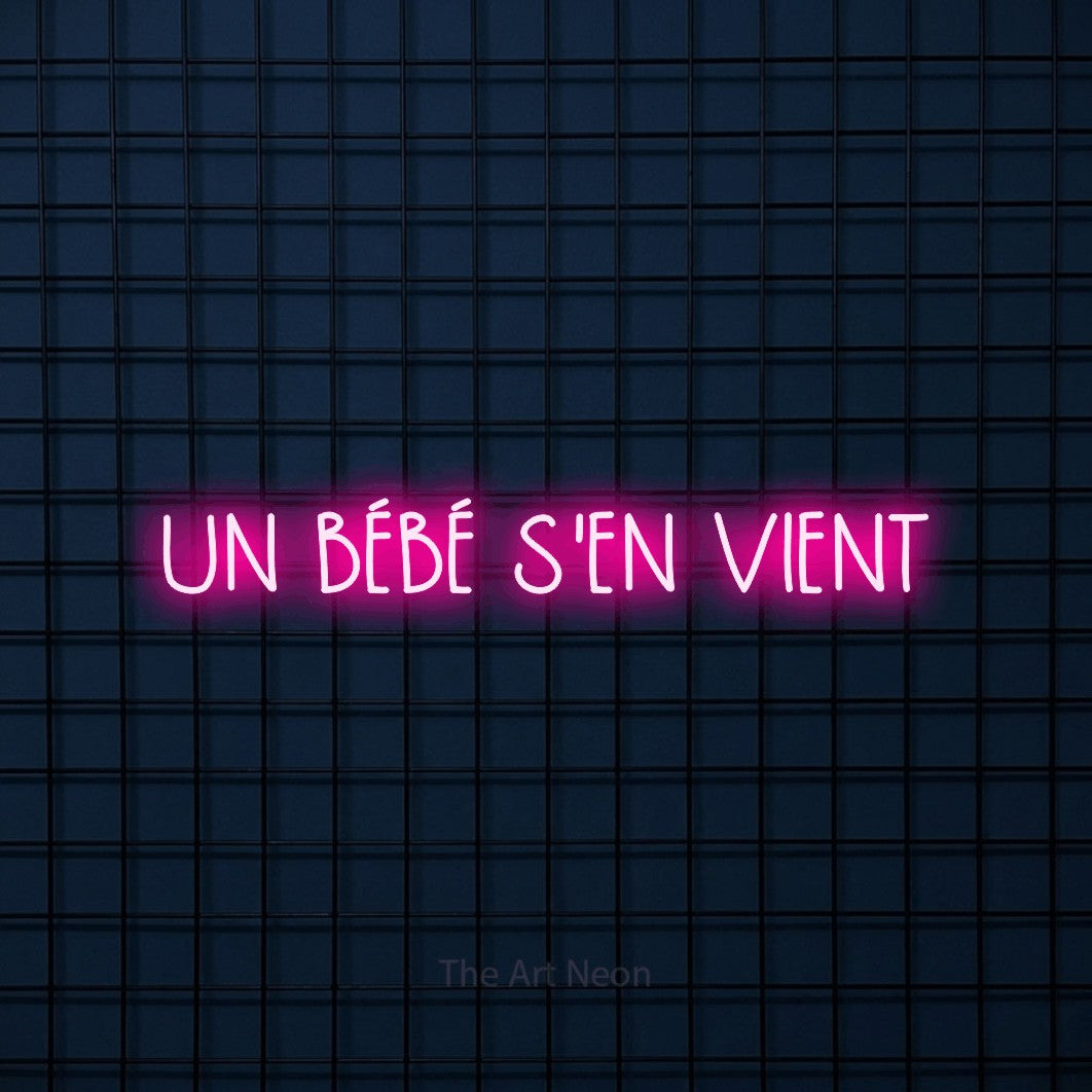 “Un bébé s'en vient” LED neon sign in hot pink, with narrow hand-drawn lettering, shown against a dark blue grid.