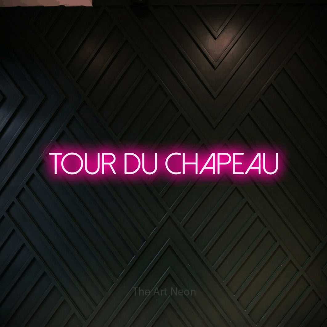 “Tour du chapeau” LED neon sign in hot pink, with thin sans-serif lettering, shown against a black chevron-patterned wood wall.