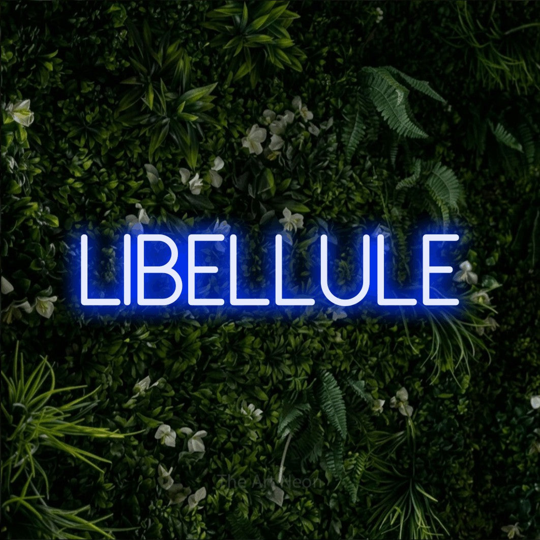 “Libellule” LED neon sign in blue, with thin sans-serif lettering, shown against a dense green plant wall.