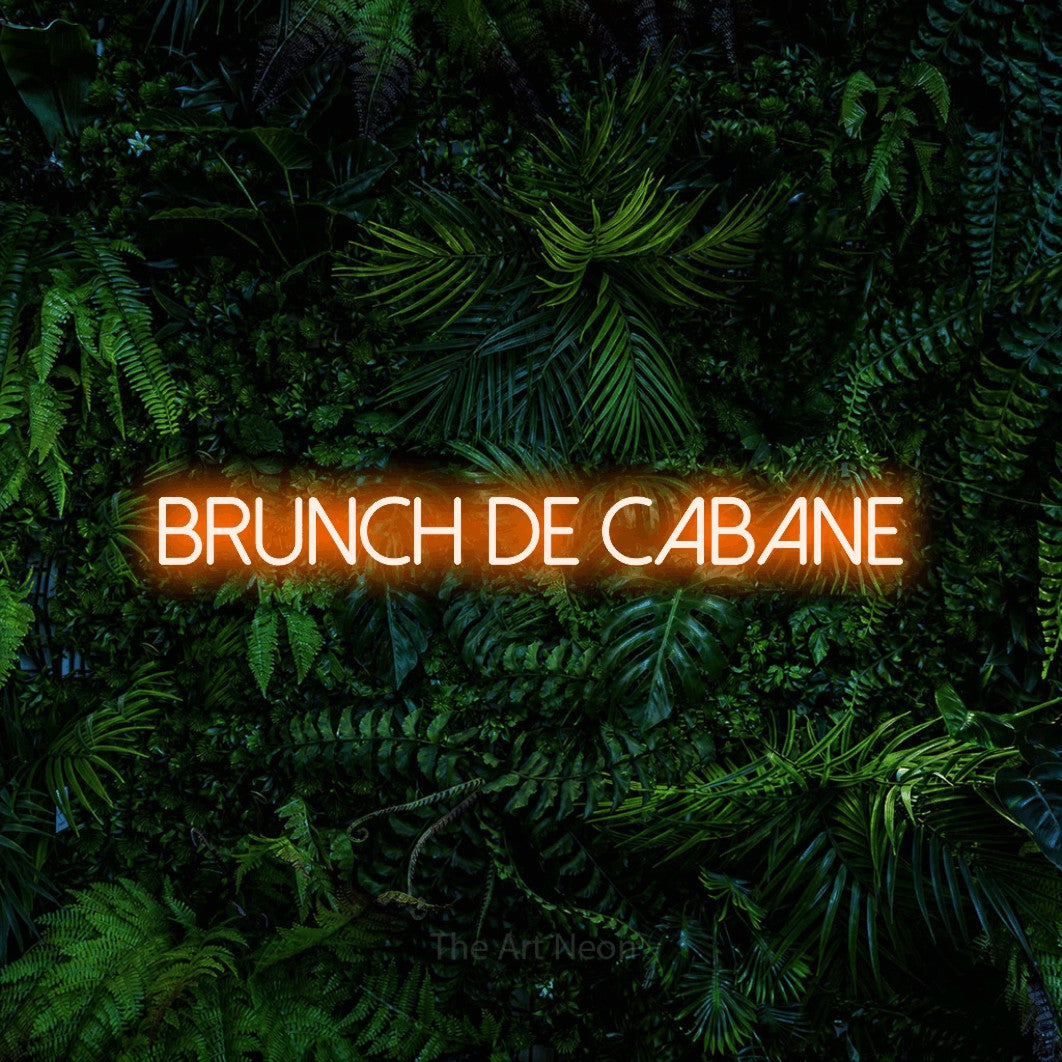 “Brunch de cabane” LED neon sign in orange, with thin sans-serif lettering, shown against dense tropical foliage.