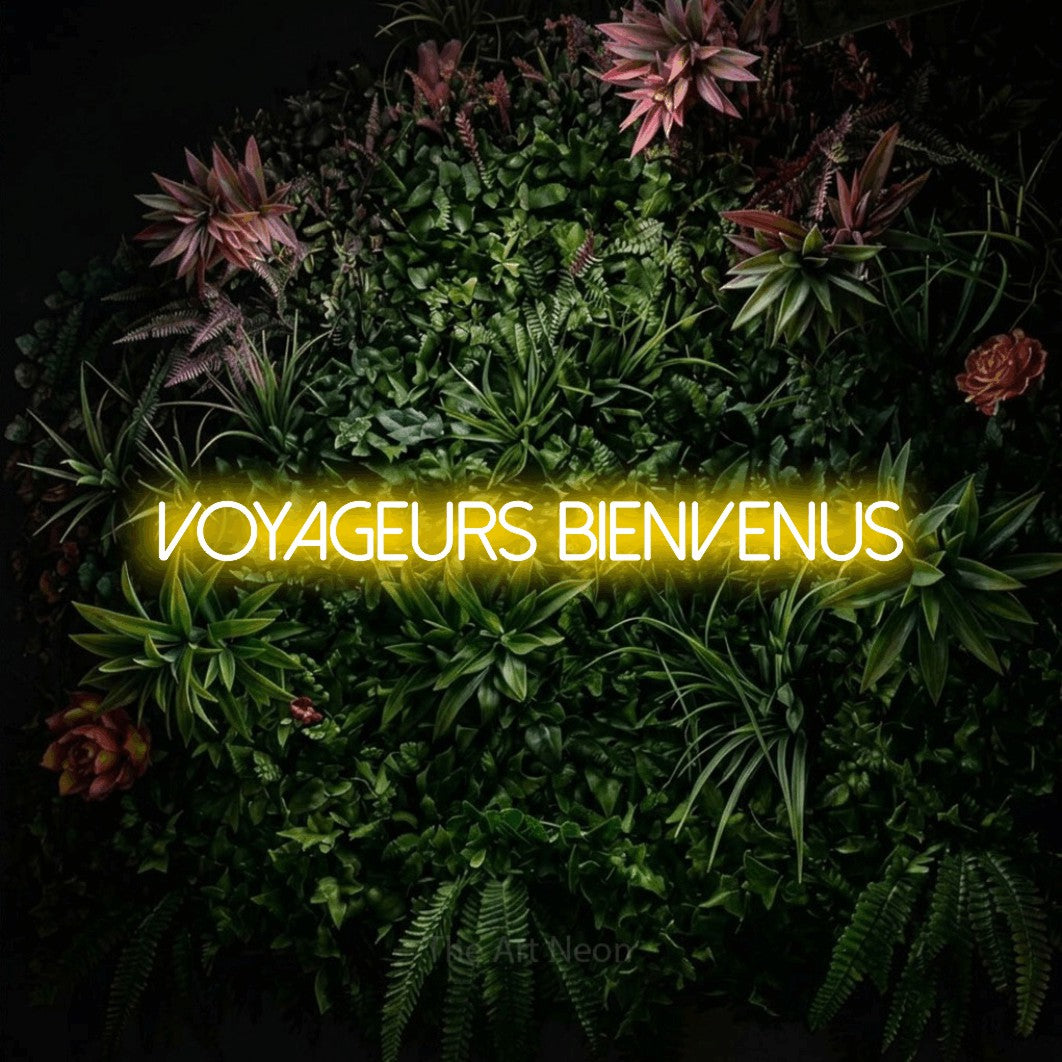 “Voyageurs bienvenus” LED neon sign in yellow, with thin sans-serif lettering, shown against a flowering plant wall.