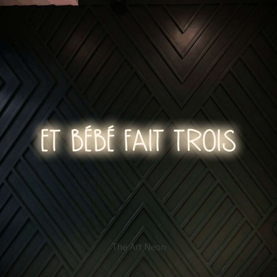 Lettrage en néon LED « Et bébé fait trois » en blanc chaud, en lettres étroites dessinées à la main, présenté sur un mur en bois noir à motif chevron, pour une fête prénatale.