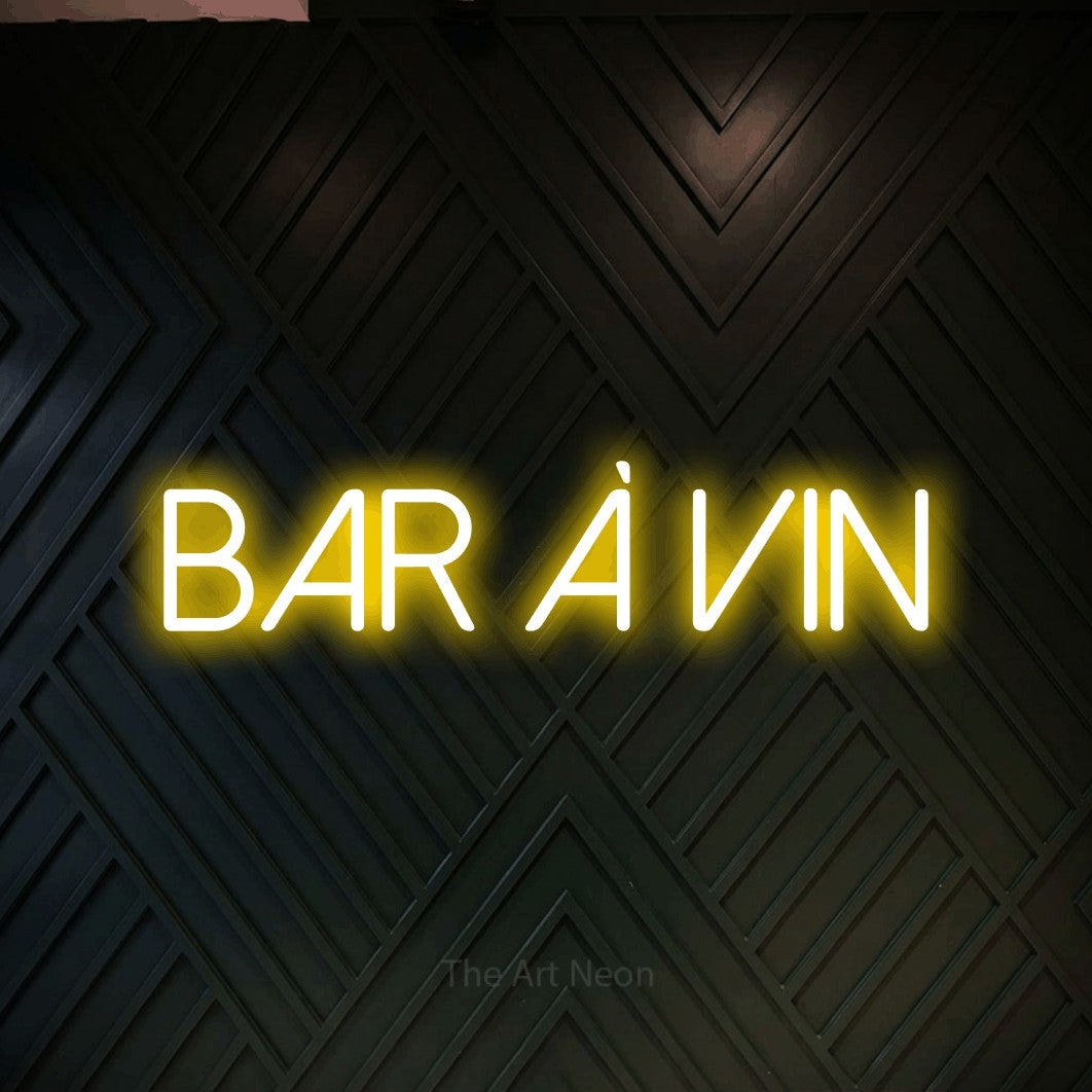 “Bar à vin” LED neon sign in yellow, with thin sans-serif lettering, shown against a black chevron-patterned wood wall.