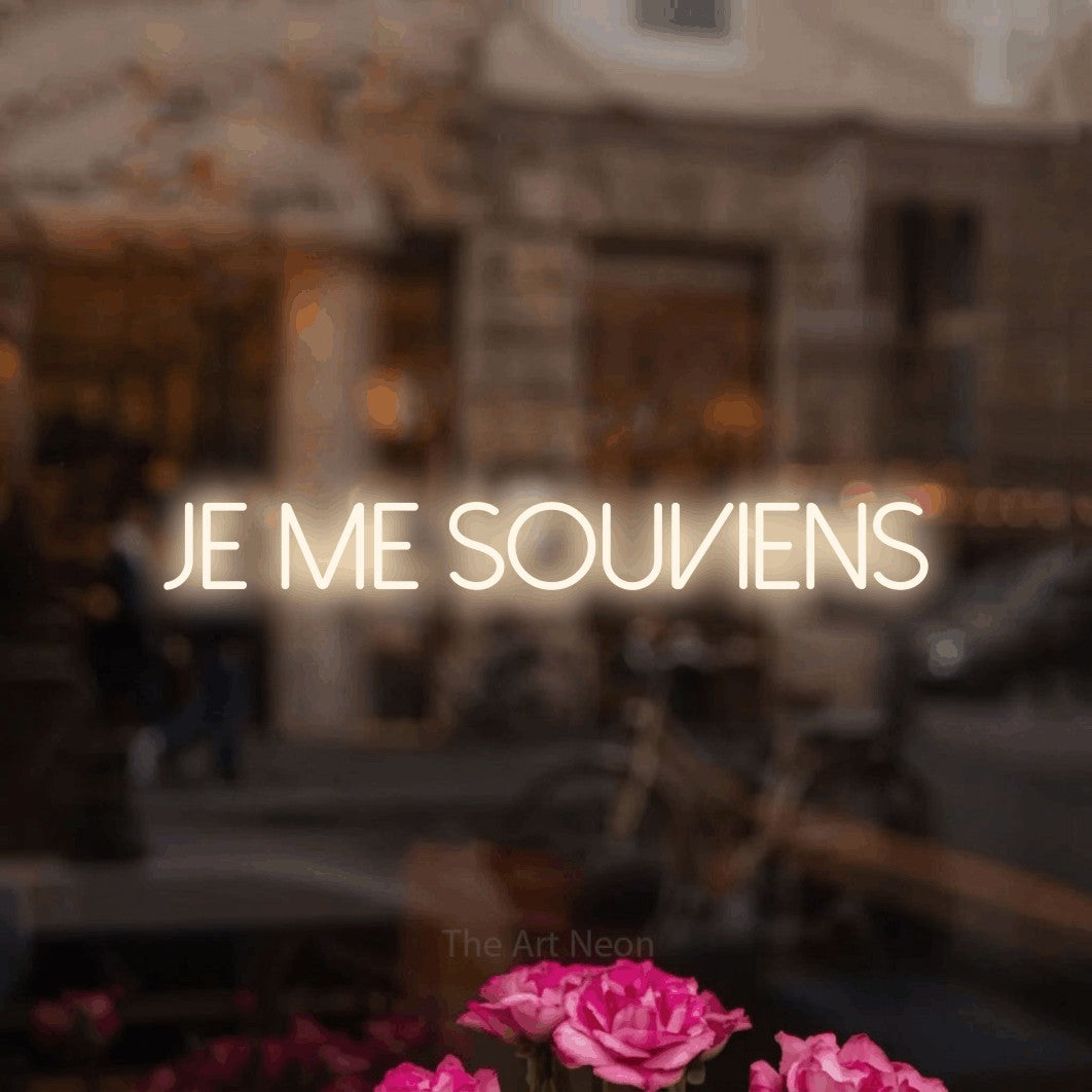 “Je me souviens” LED neon sign in warm white, with thin sans-serif lettering, shown against a blurred café-bar interior.